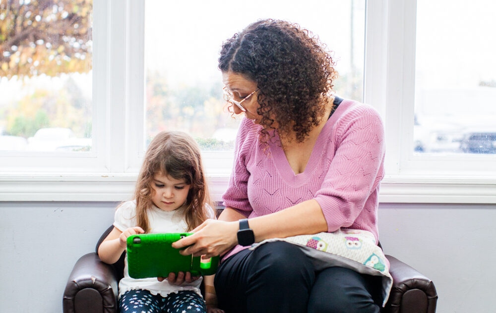 Child using tablet held by adult while they're sitting on a couch