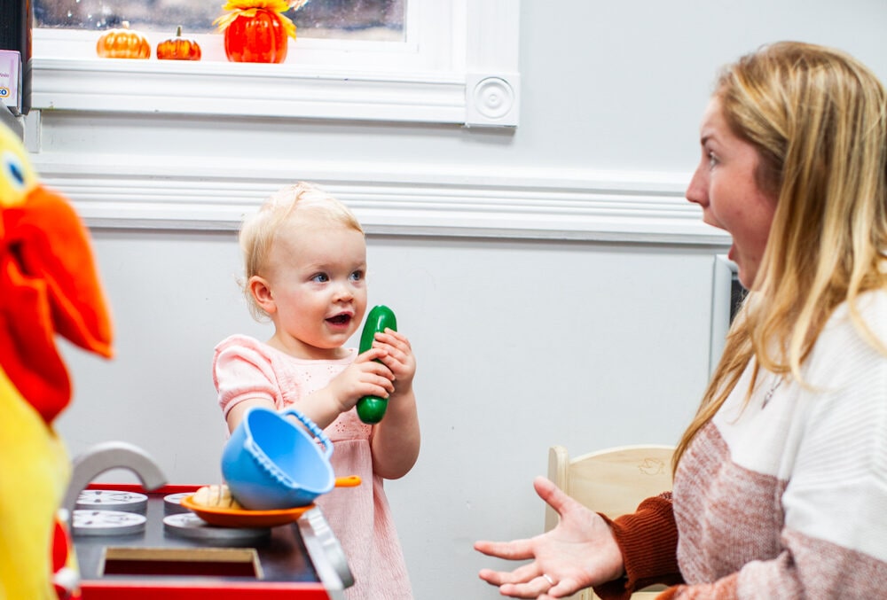 Toddler holding plastic cucumber toy and showing it to surprised teacher